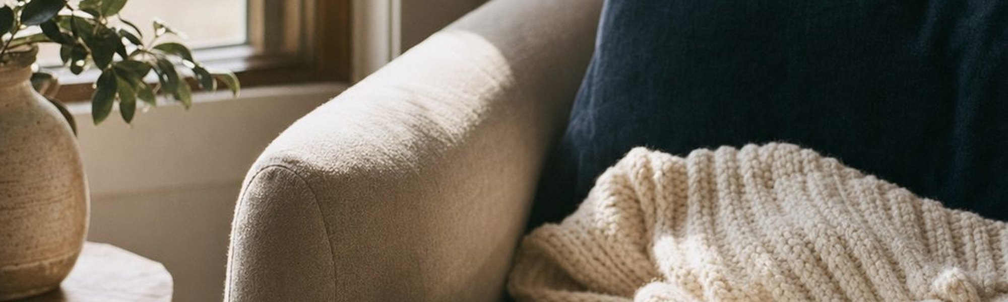 A comfortable armchair by a sunlit window with a knit blanket and a plant.